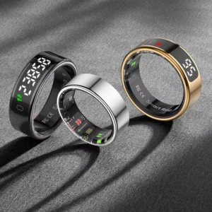 Three smart rings in black, silver, and gold finishes, resting on a dark textured surface. The black and gold rings feature a digital display on the outer band.